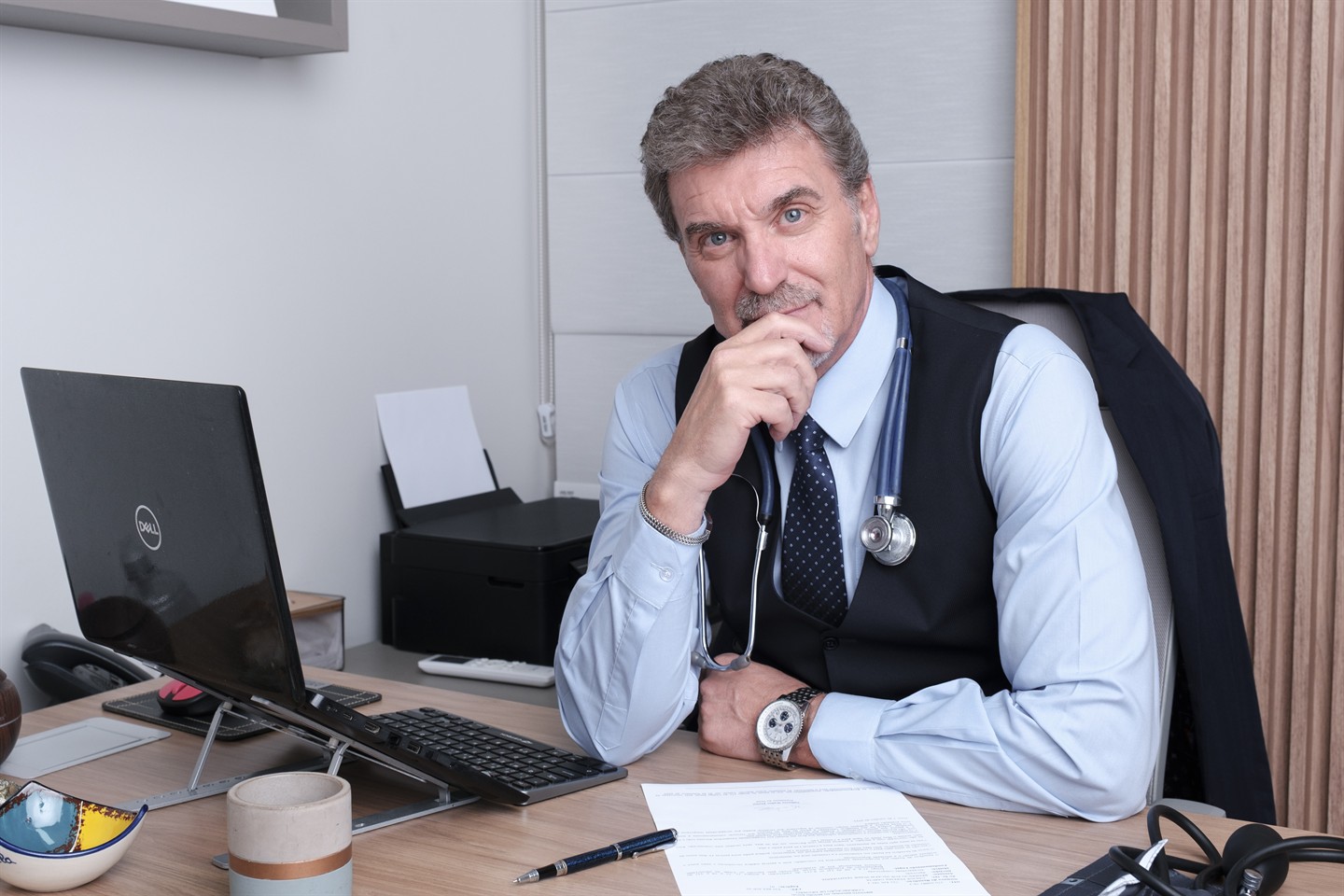 Médico em consultório, sentado à mesa com estetoscópio.